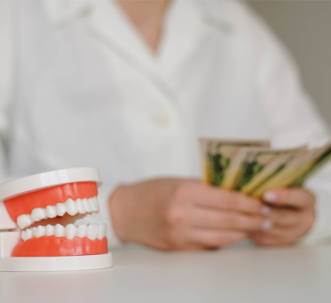 Dentist in white coat holds dental model showing teeth while reviewing patient chart at desk.