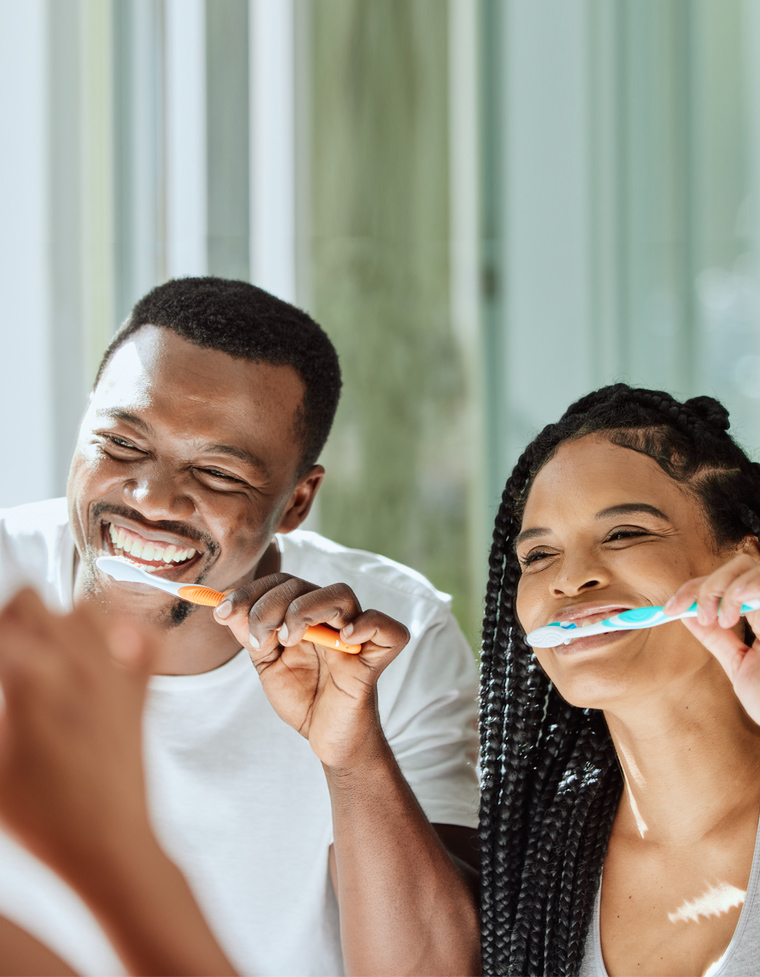 Man and woman brushing teeth together in bathroom, smiling at camera.